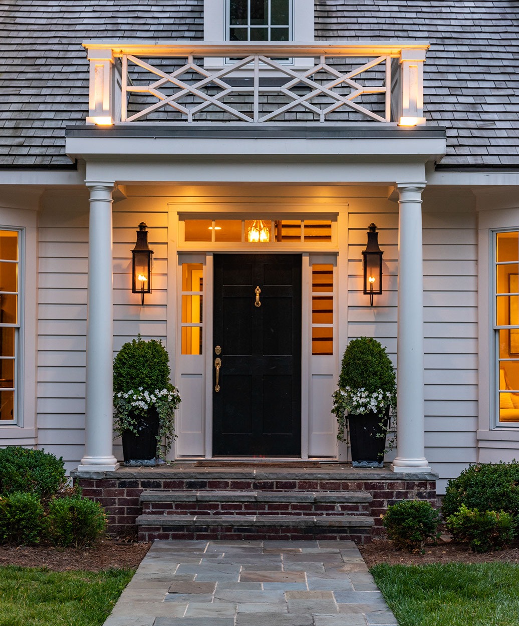 Front view of a welcoming house entrance with a black door, white trim, two potted plants, brick steps, and warm lights glowing from wall lanterns and windows on both sides. A small balcony is visible above the porch.