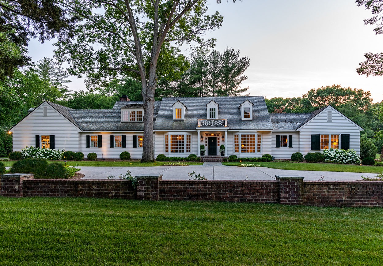 A large, white, two-story house with a gray roof, black shutters, and dormer windows, surrounded by trees and landscaping, seen at dusk from across a grassy lawn and brick wall.