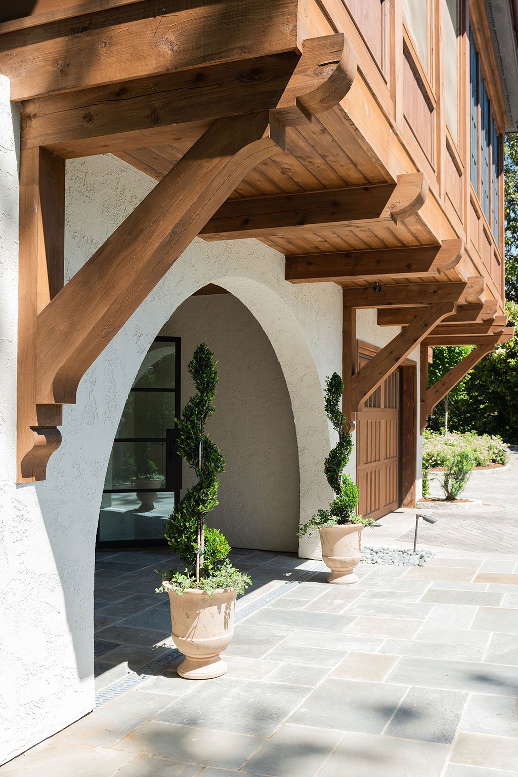 Sunlit entryway of a house with a white stucco wall, wooden beams, arched entry, and two potted spiral topiary plants on a stone patio.