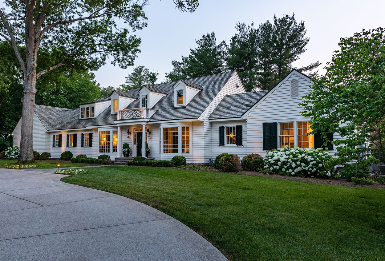 A large white house with a gray roof, black shutters, dormer windows, and a manicured lawn with shrubs and flowers, seen at dusk with interior lights glowing. A curved driveway leads to the entrance.