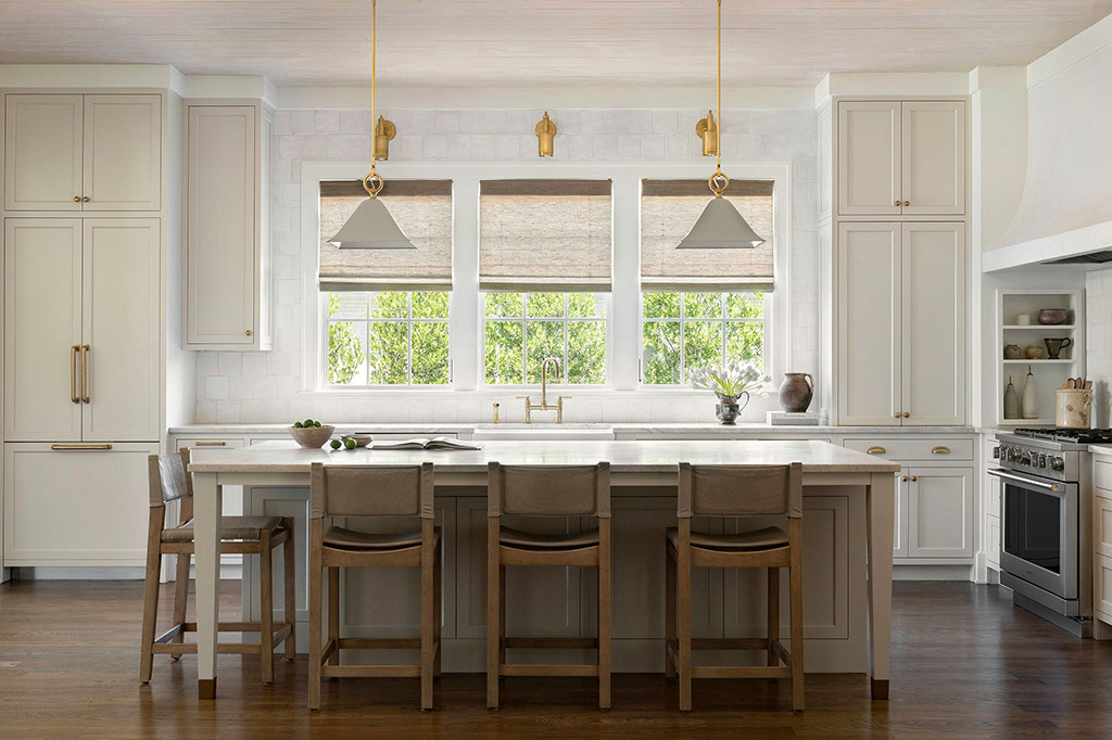A modern, spacious kitchen with white cabinets, gold hardware, a large island with four wooden barstools, pendant lights, wide windows with shades, and stainless steel appliances.