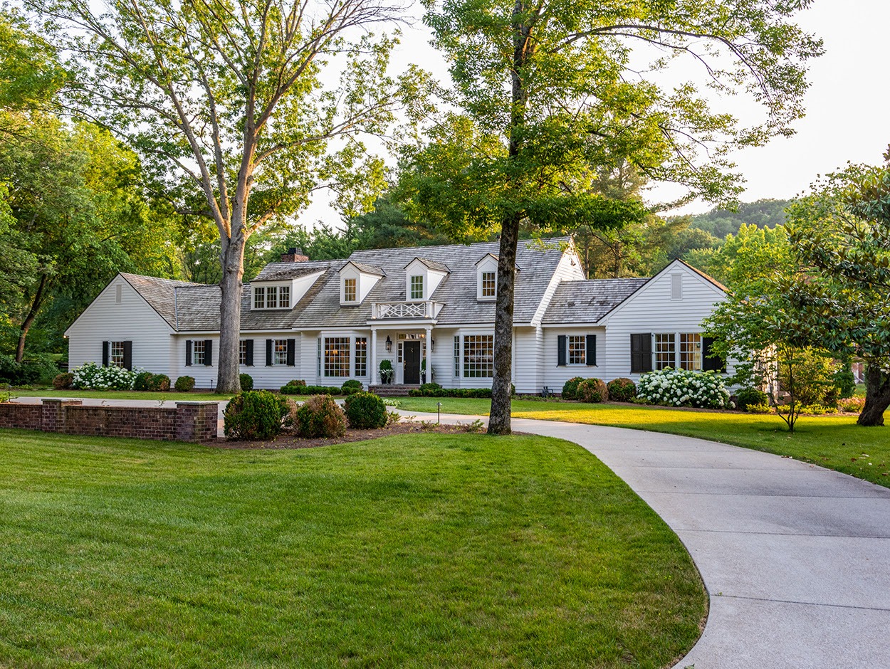 Large white two-story house with gray shutters and a shingled roof, surrounded by lush green lawn, manicured bushes, and tall trees. A curved concrete driveway leads up to the front entrance.