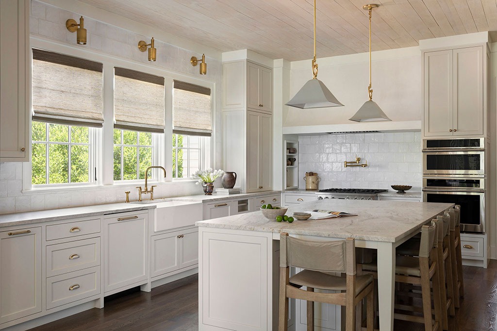 Bright, modern kitchen with white cabinets, marble countertops, a large island with wooden stools, gold fixtures, pendant lights, farmhouse sink, large windows with blinds, and built-in appliances against a white tile backsplash.