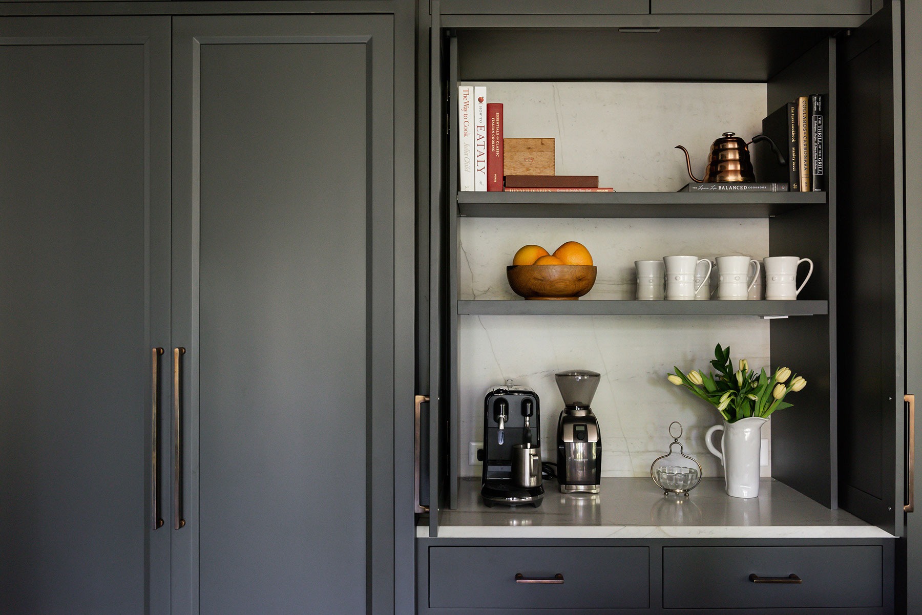 A kitchen nook with gray cabinets, a coffee machine, grinder, a bowl of oranges, stacked cups, books, a teapot, a vase with white tulips, and drawers with brass handles.