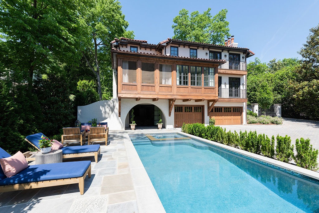 A luxurious house with a tiled roof and wooden balcony overlooks a rectangular swimming pool, surrounded by lounge chairs, trees, and neatly landscaped greenery under a clear blue sky.