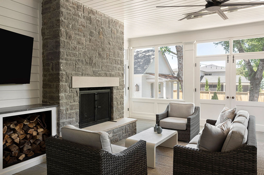 A modern sunroom with wicker furniture, a stone fireplace, a mounted TV, and stacked firewood. Large windows and glass doors let in natural light, offering a view of a yard and neighboring house.