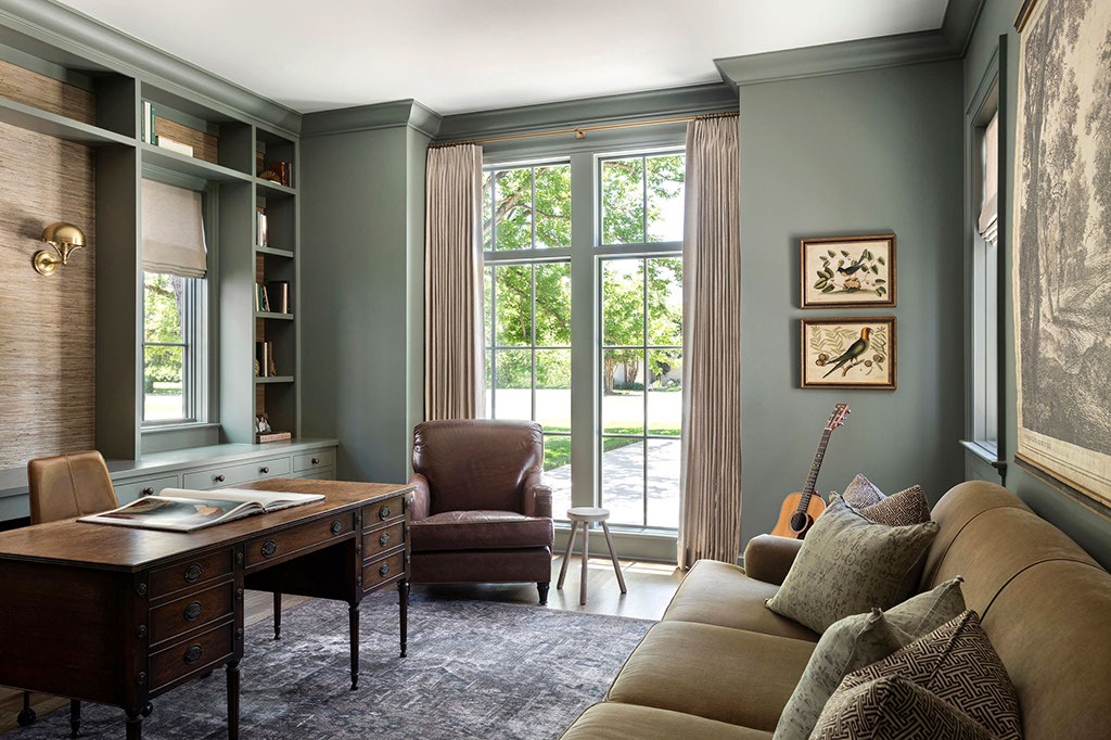 A cozy home office with a vintage wooden desk, leather armchair, beige sofa, bookshelves, and large window letting in natural light. Bird artwork and a guitar decorate the room. Neutral greenish-gray walls and soft furnishings.
