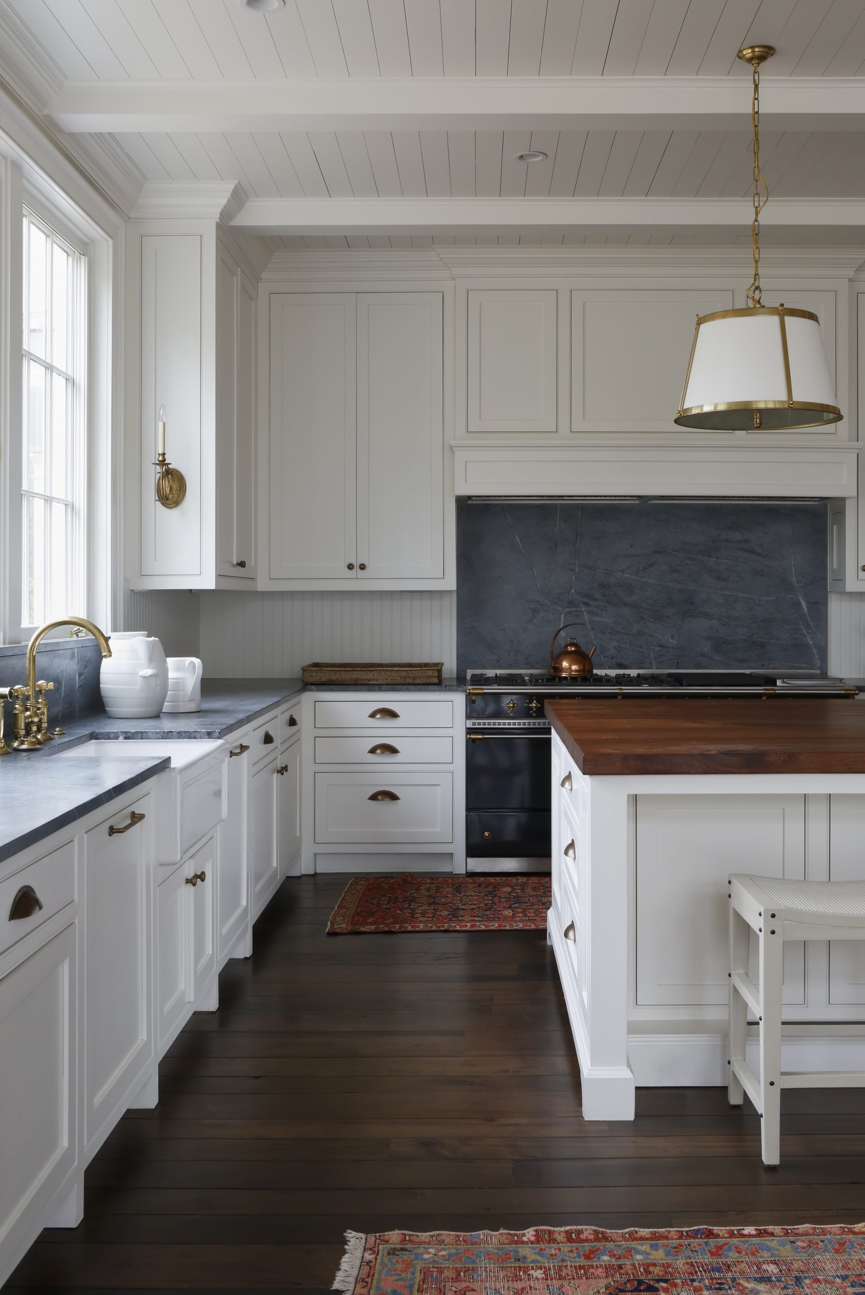 A modern kitchen with white cabinets, dark countertops, and a wooden island. The space features brass fixtures, pendant lighting, a dark range, and decorative rugs on dark wood floors. Large windows provide natural light.