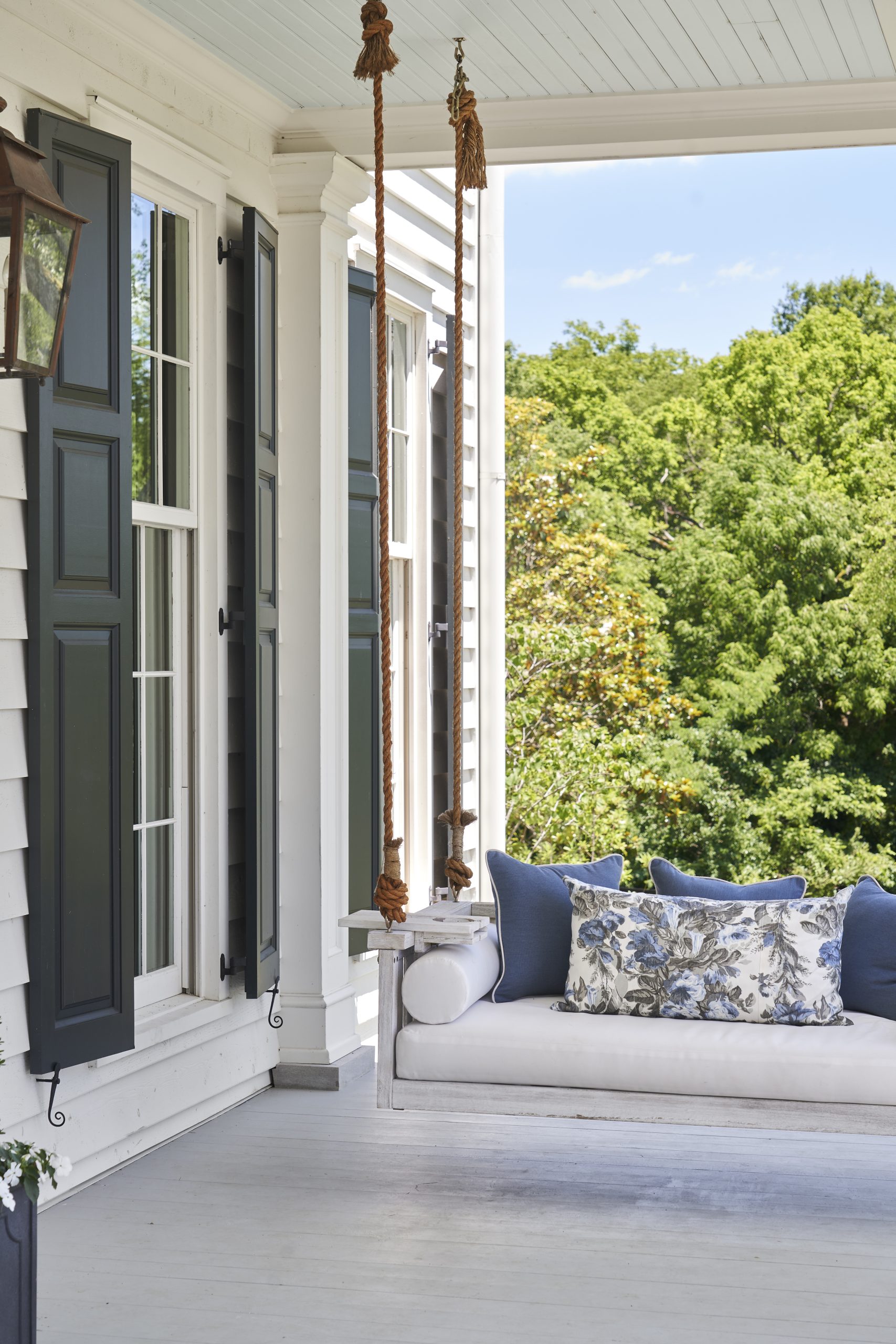 A cozy porch swing with blue and white pillows hangs from ropes on a white-painted porch, next to tall windows with dark shutters, overlooking lush green trees on a sunny day.
