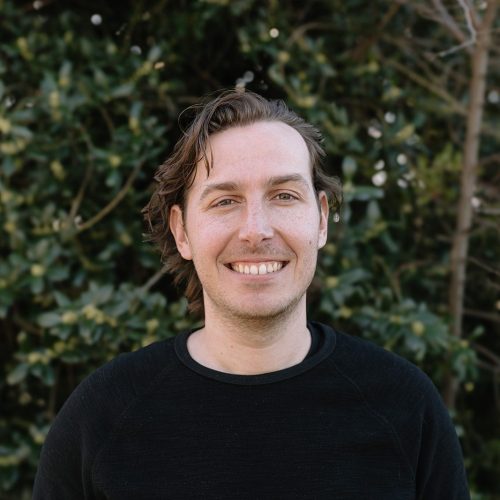 Rankin Designworks A man with light skin and brown hair smiles at the camera, standing outdoors in front of green leafy trees. He is wearing a black sweater.