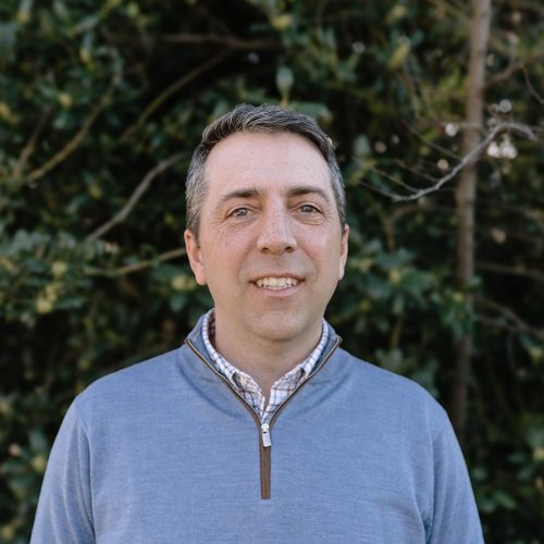 Rankin Designworks A man with short brown hair, wearing a light blue quarter-zip sweater over a plaid shirt, stands outdoors in front of leafy green bushes, smiling at the camera.